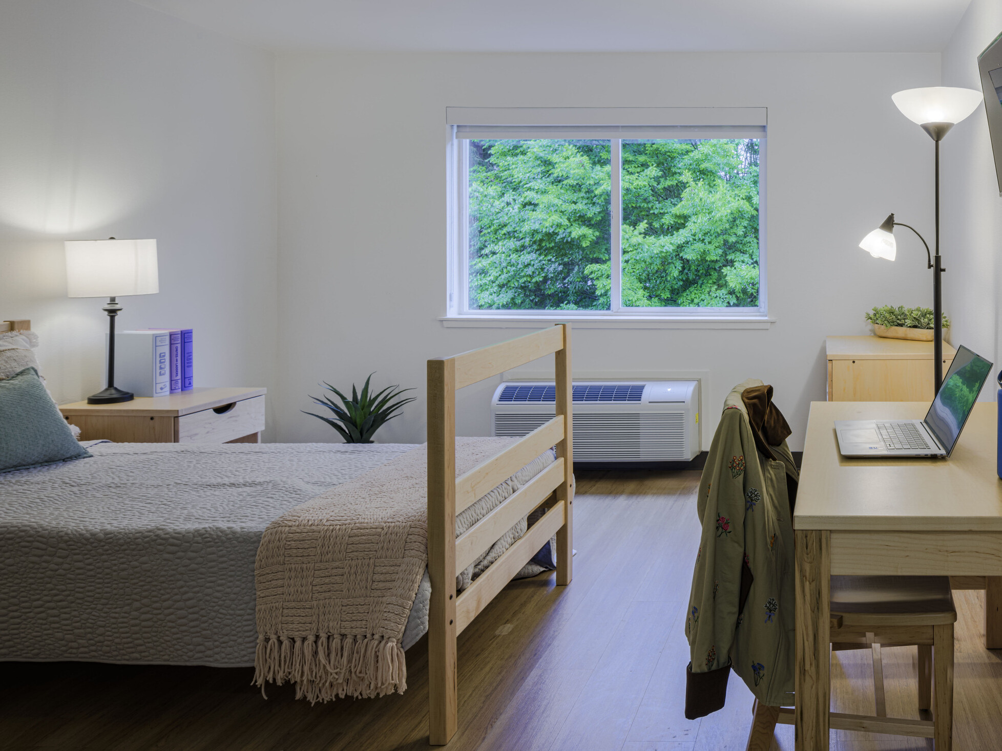 A resident room contains natural wood furniture, a bed with a beige throw, a compact desk with a laptop and chair, wall-mounted TV, and a window overlooking greenery