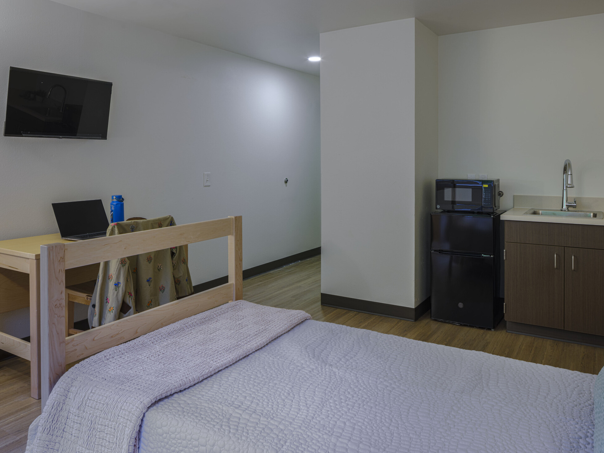 From the bed in a resident room, the view includes a kitchenette equipped with a mini-fridge, microwave, and sink, as well as a workspace with a desk, laptop, and wall-mounted monitor