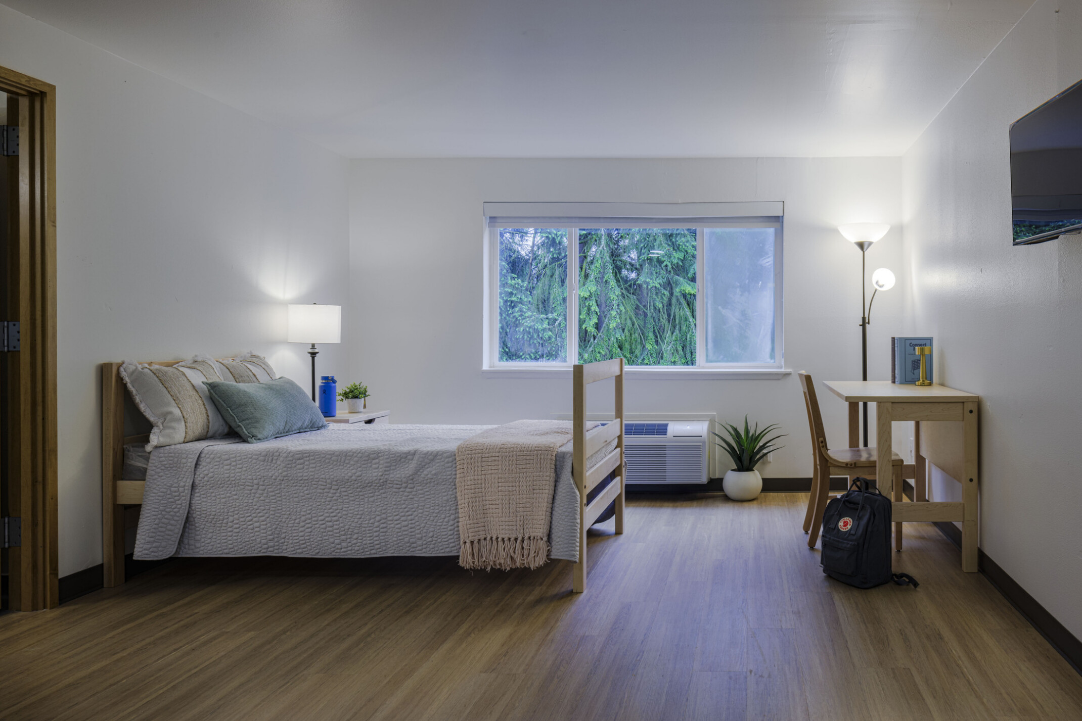 A dormitory-style room features a neatly made bed, compact desk, and a window on the far wall, with neutral colors and varied textures.