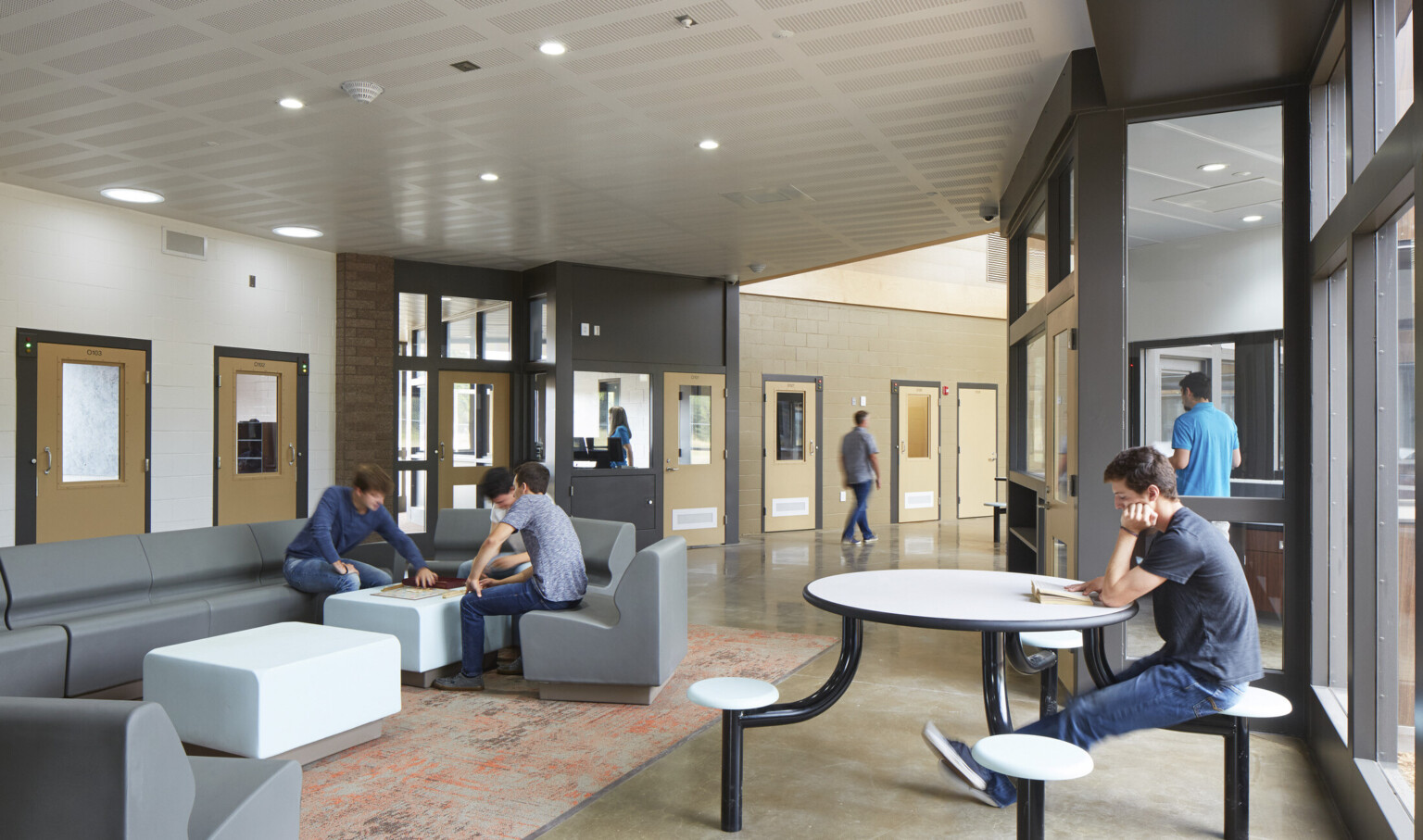 Partially carpeted seating area with tables in grey and white room at the end of white hallway. Light wooden doors line hall