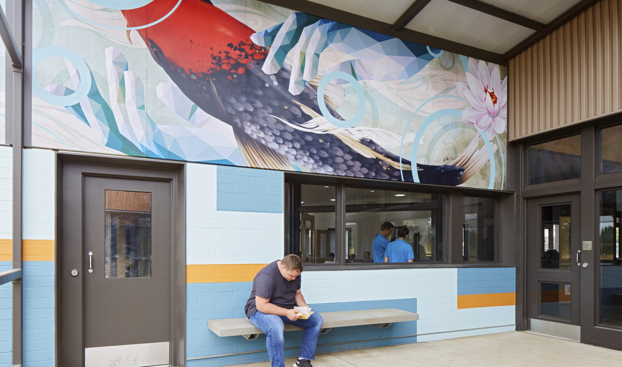 A grey bench against a blue and yellow colorblock wall with a window into an office. Above is a mural of a koi fish