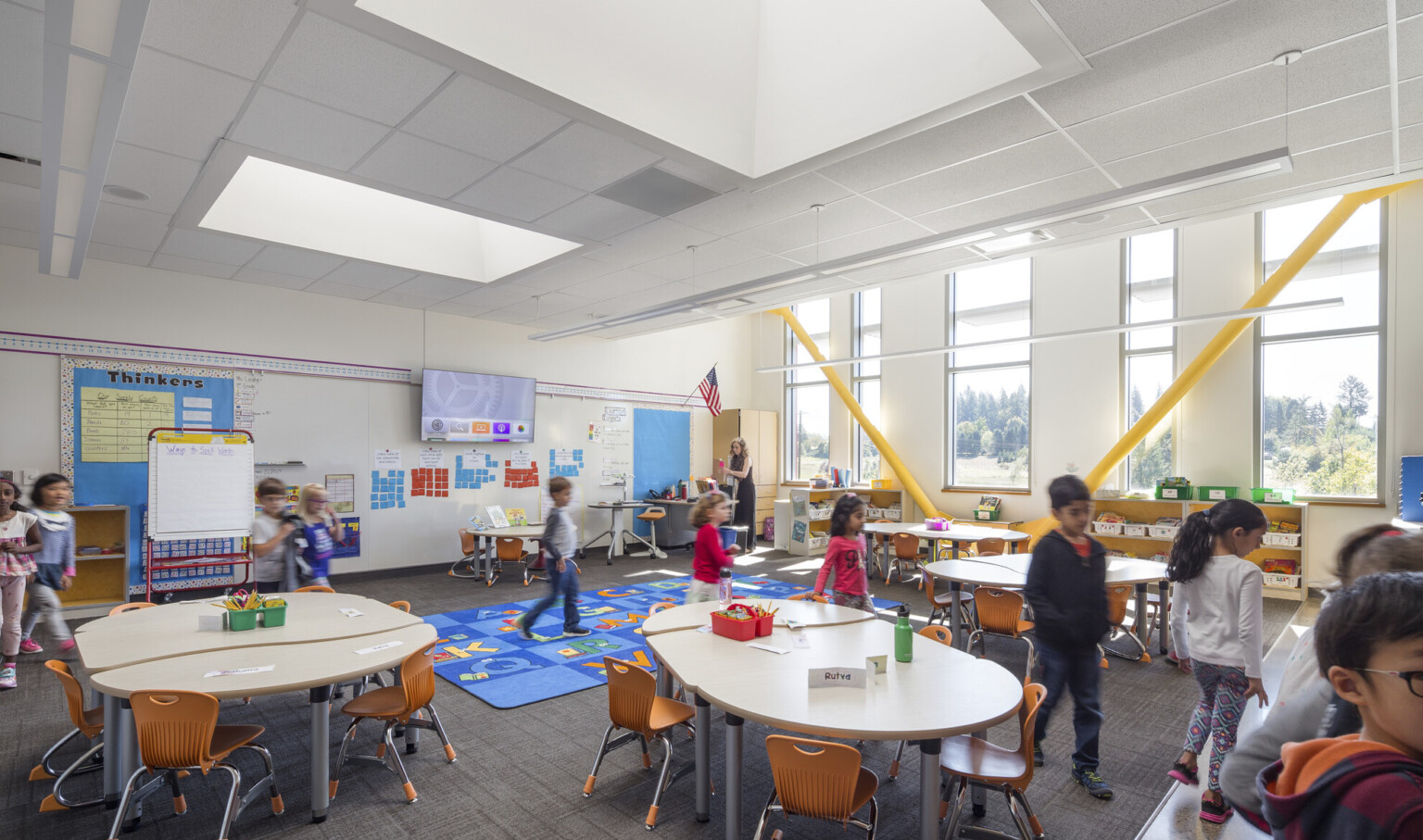 Classroom with yellow exposed beams at angles by windows, right. Oval kid sized tables and chairs around blue alphabet carpet