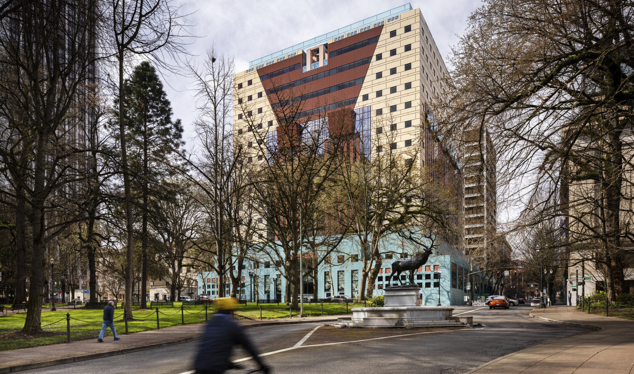 View of beige building with red angular stripe, blue ribbon design at side, over a teal multistory base, surrounded by trees