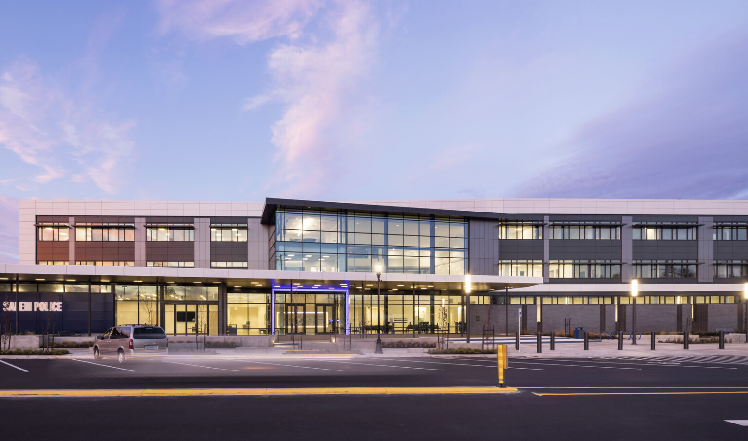 Front view of police station from across parking lot with angled overhang, double height windows, and entry lit in blue