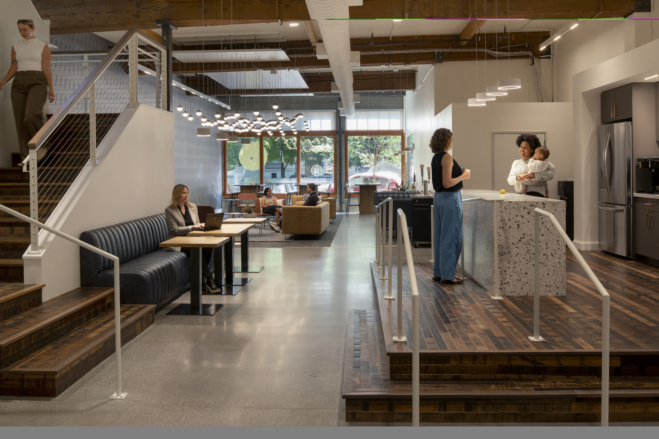 Work café with kitchen island where two employees stand talking, one holding a baby; a staircase to one side and varied seating where people work solo and in groups