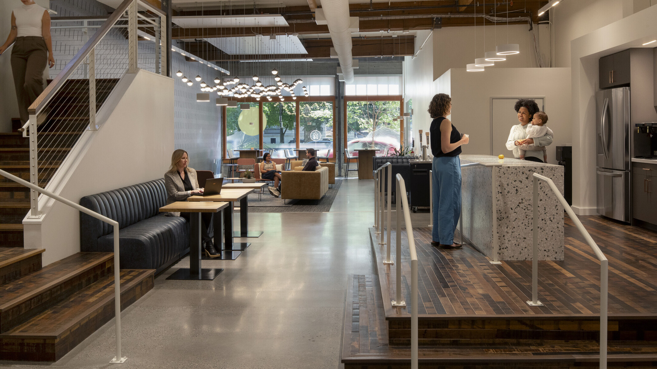Work café with kitchen island where two employees stand talking, one holding a baby; a staircase to one side and varied seating where people work solo and in groups