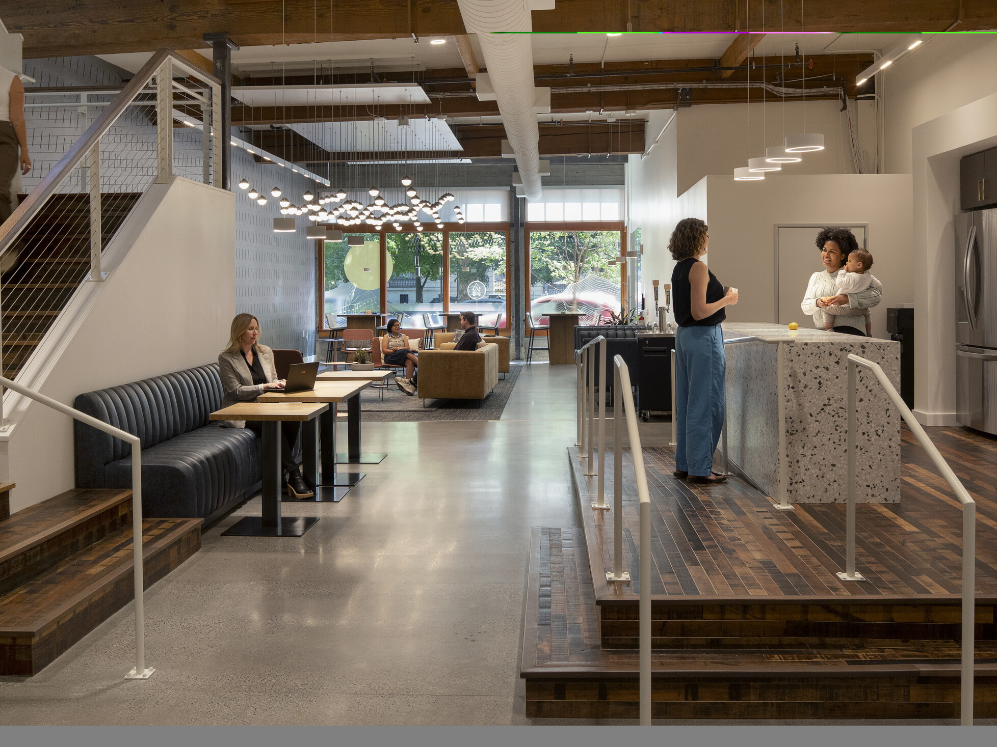 Work café with kitchen island where two employees stand talking, one holding a baby; a staircase to one side and varied seating where people work solo and in groups