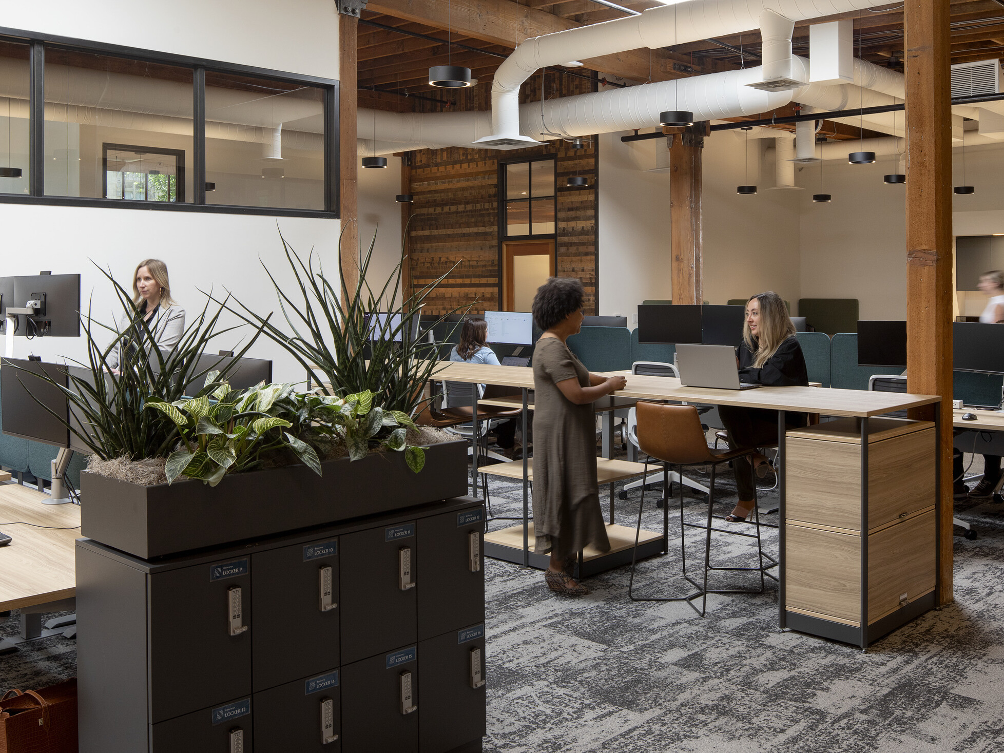Workstation area with rows of desks and long hightop tables, day-use lockers topped with planting at the end of the workstations, and exposed wood beams and columns overhead