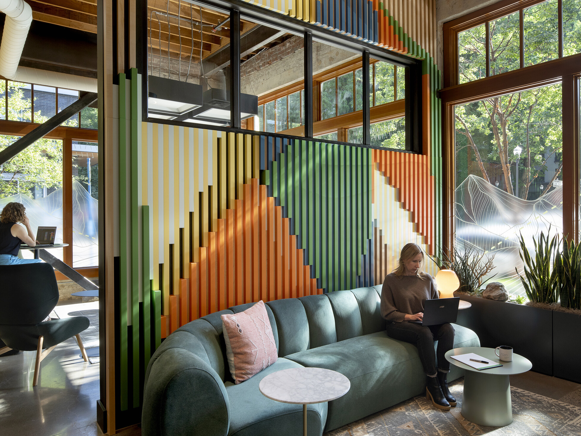 Casual working area divided by an open, doorless color wall in orange, green, gold, and blue; a woman works on her laptop on a green couch on one side, while someone works at a high top table on the other