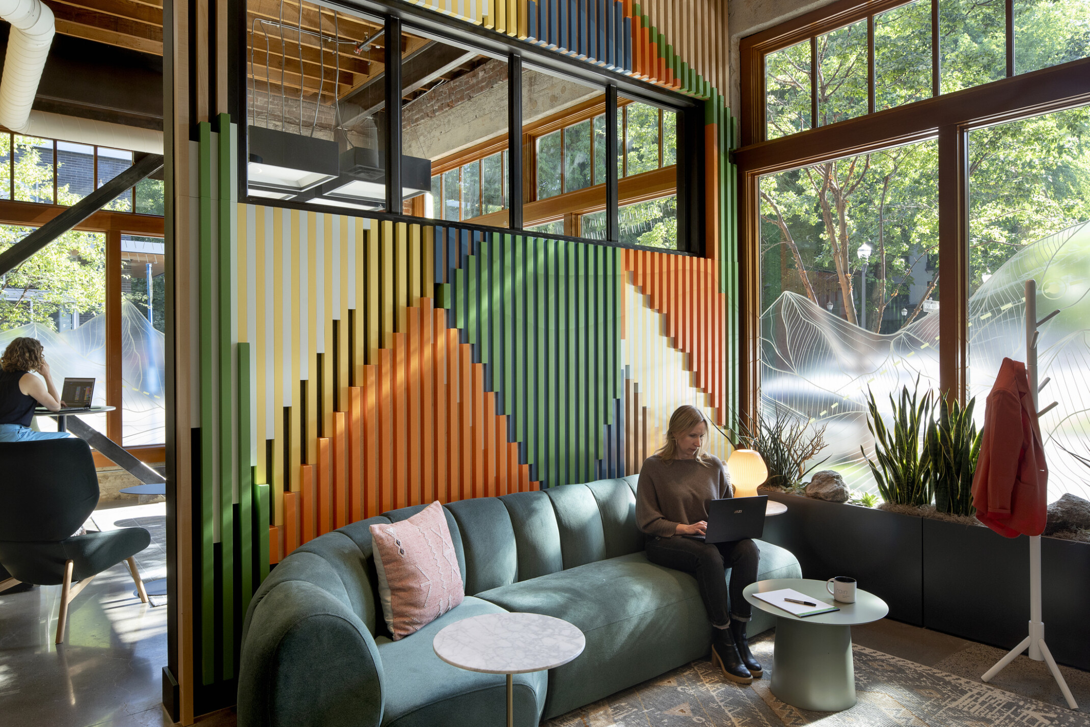 Lady sitting on a green couch working on a computer with a orange, green and blue wall behind the couch