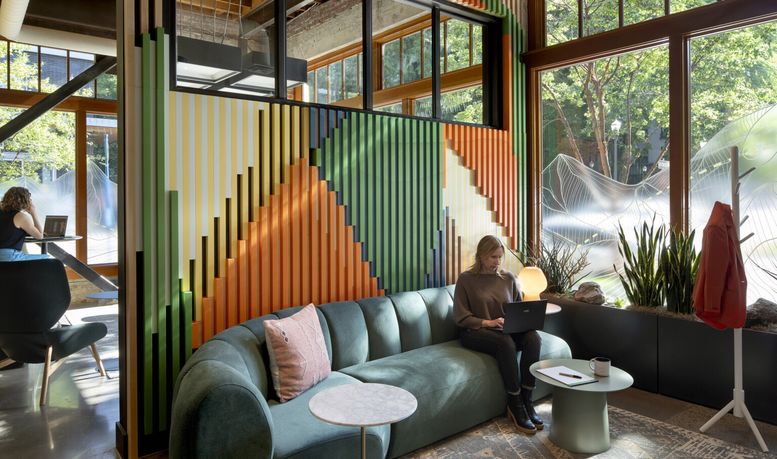 Lady sitting on a green couch working on a computer with a orange, green and blue wall behind the couch