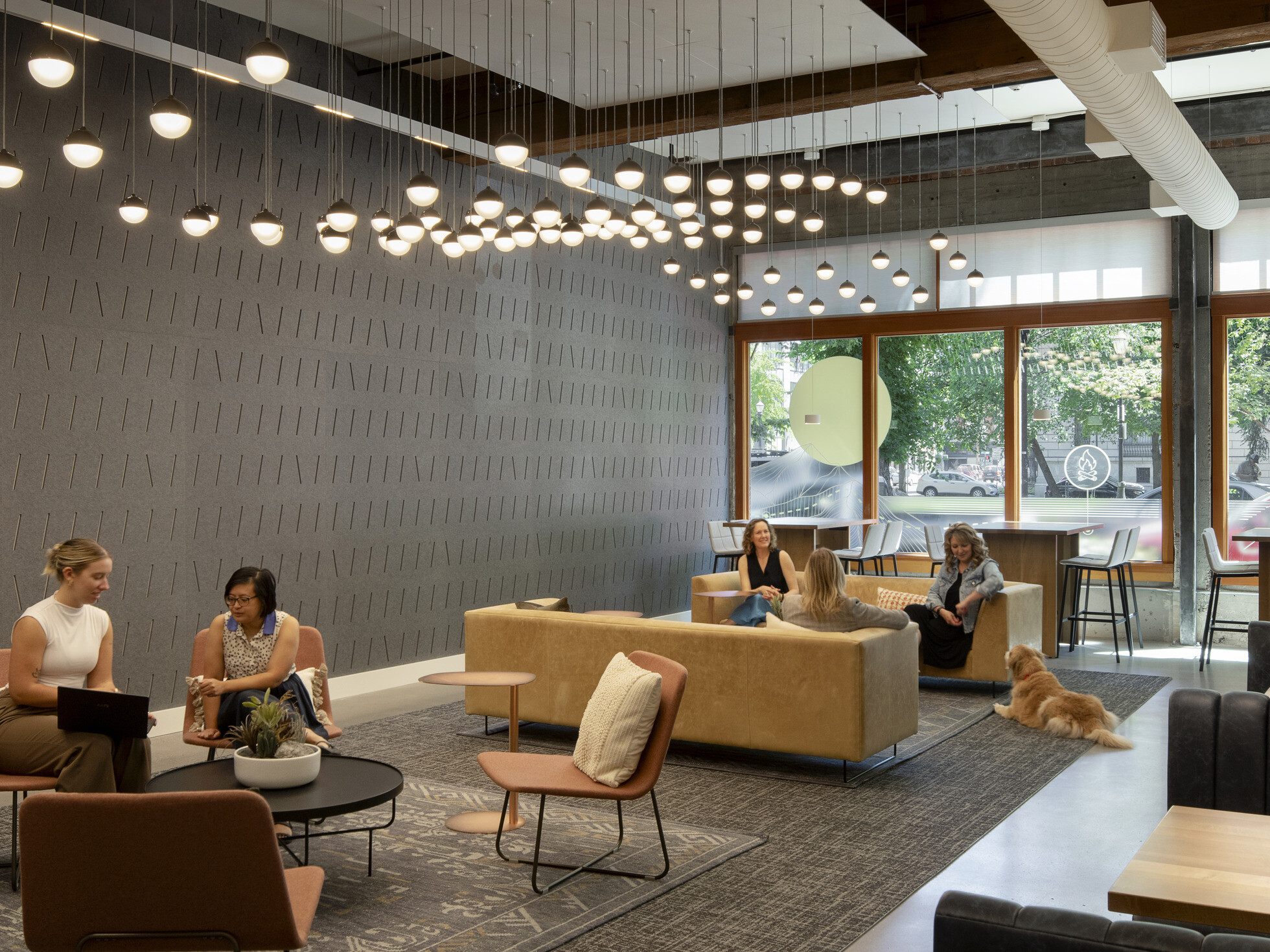Casual seating area connected to the work café with couches, single chairs, coffee tables, and booths; groups of people chat together, including one with a Labrador retriever