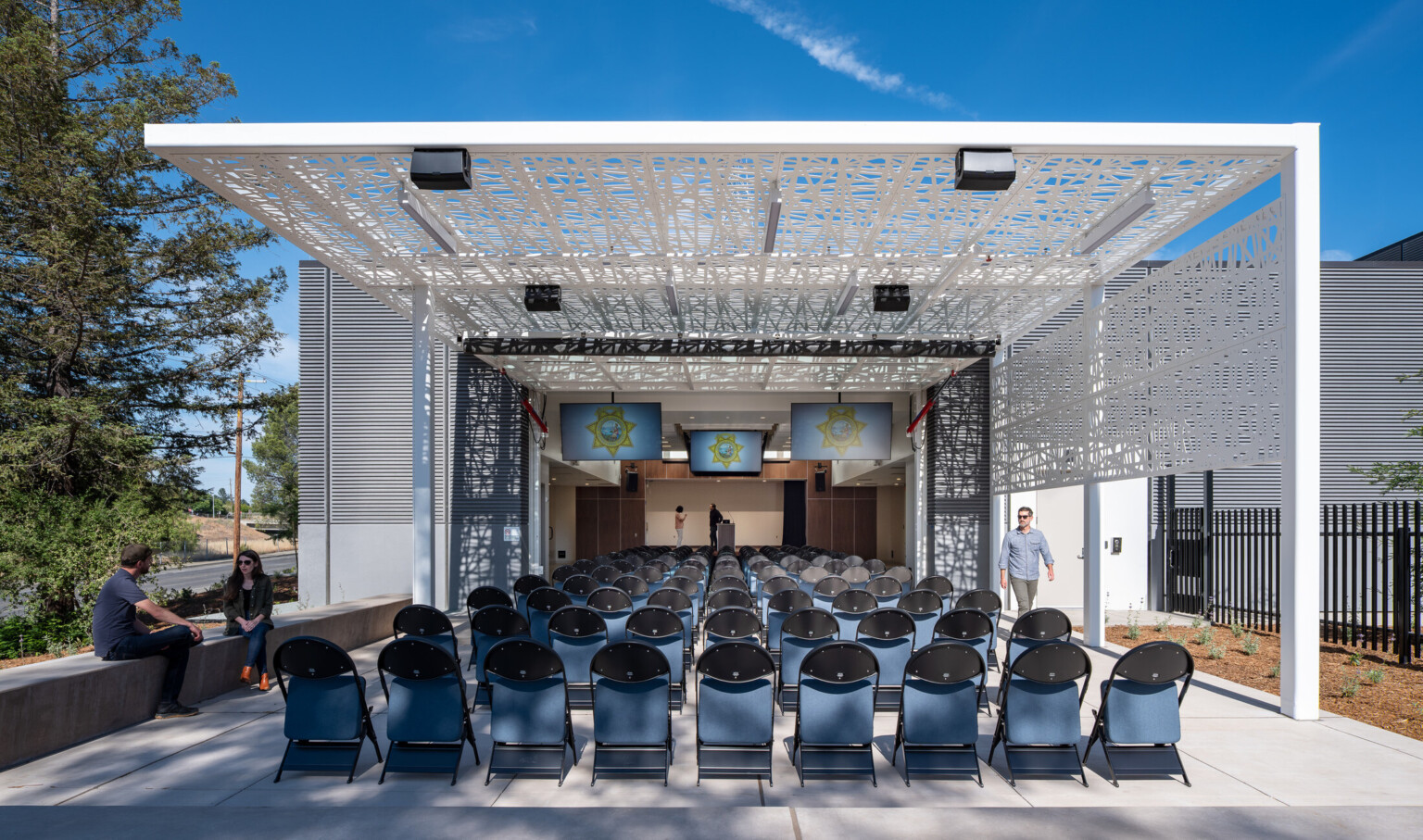 Exterior courtyard filled with chairs covered by a modern white awning looking at a podium with TV's suspended from the ceiling Exterior courtyard filled with chairs covered by a modern white awning looking at a podium with TV's suspended from the ceiling