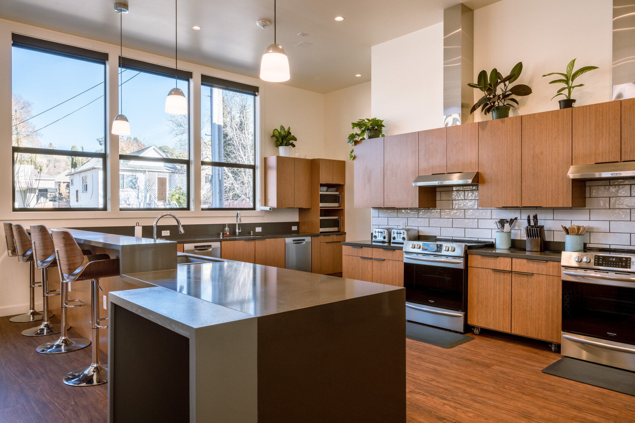 Multi-use kitchen with tan wood cabinets; peninsula counter with high top seating; three large windows in the background bringing in natural light