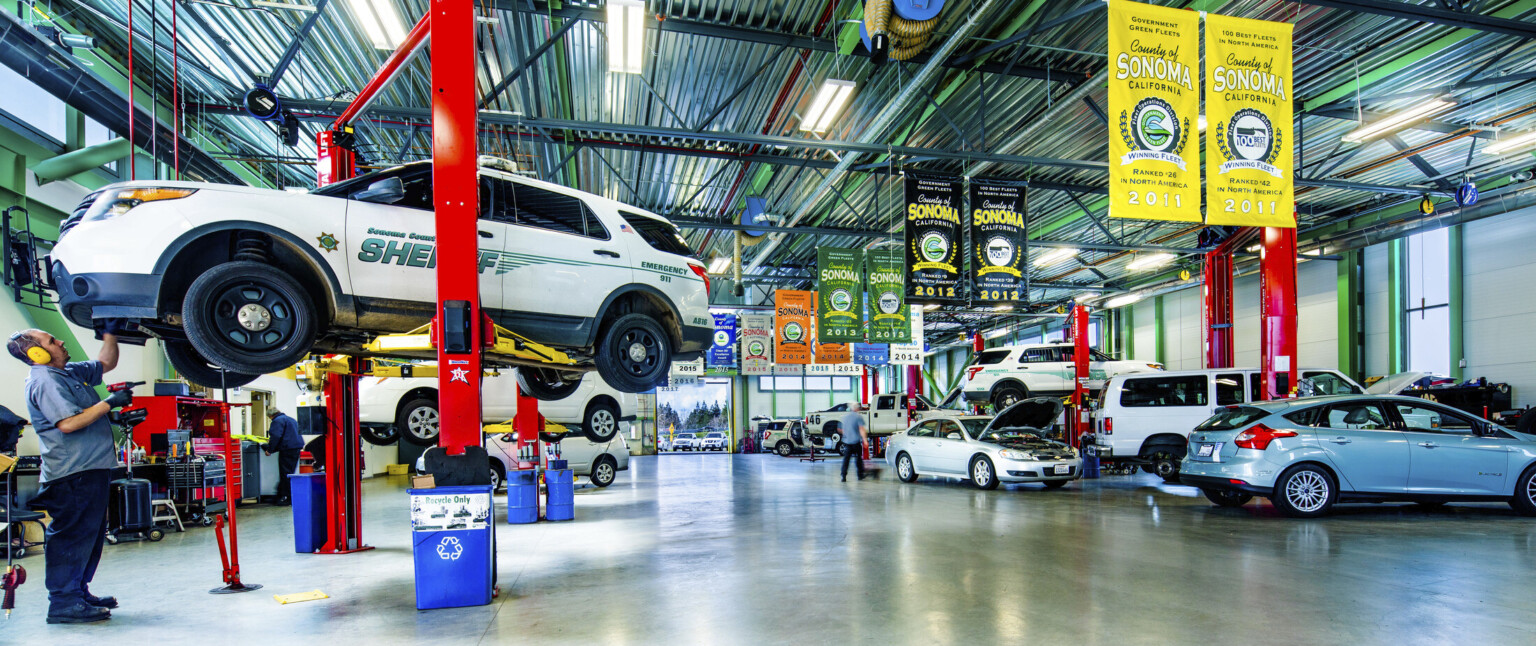 Interior with cars along side walls, some lifted by a red and yellow machine. Banners hang along center from paneled ceiling