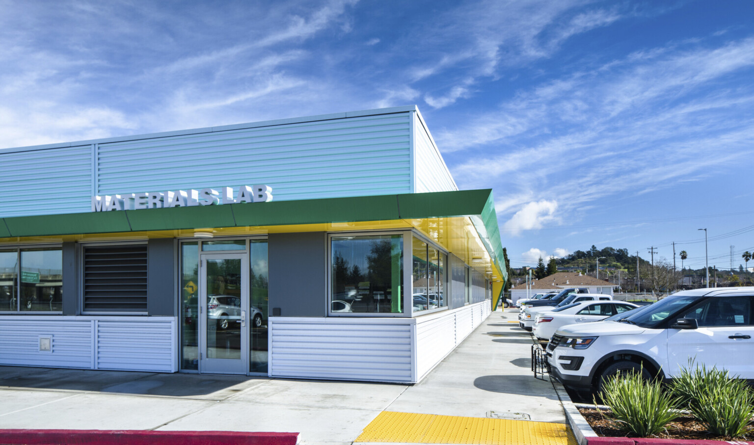 Silver panel building with green and yellow awning with sign reading Materials Lab over glass door. Parking lot to the right