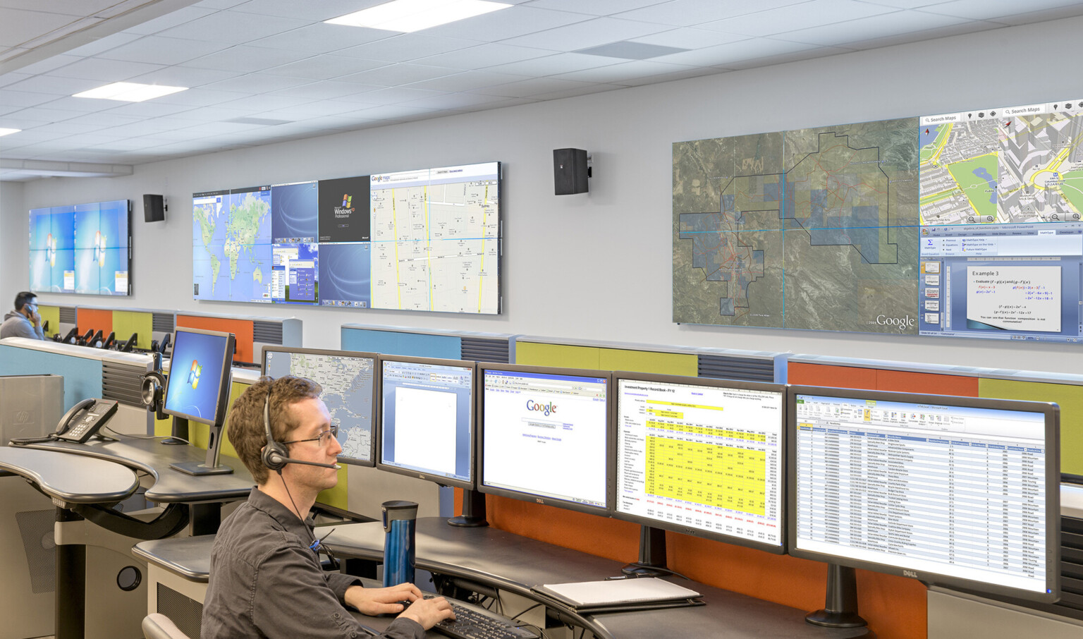 Man wearing headset sits at desk with 5 screens of spreadsheets and maps with colorful wall panels behind
