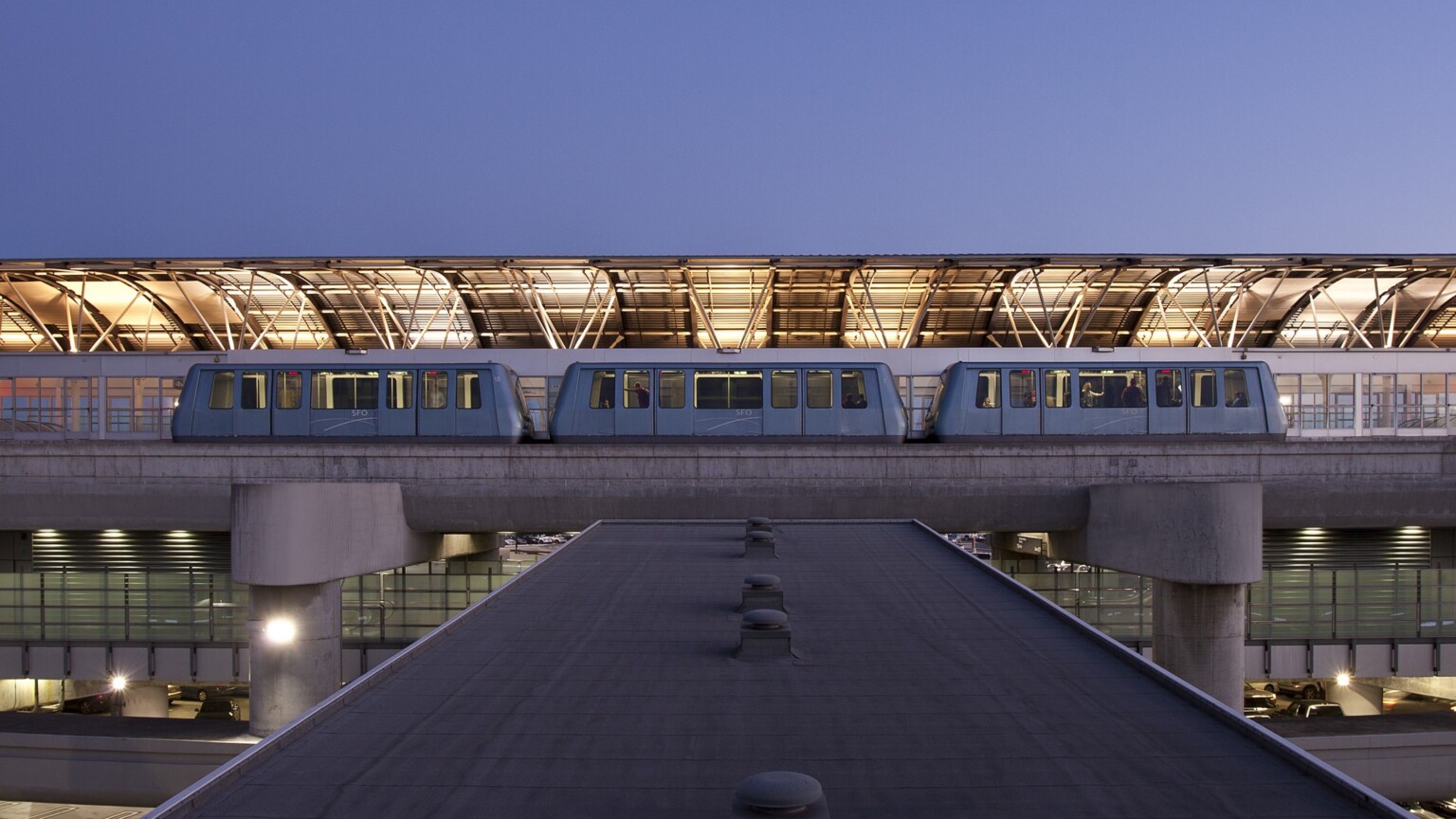View from top of skywalk to train at station with illuminated curved half roof on opposite side with exposed beams.
