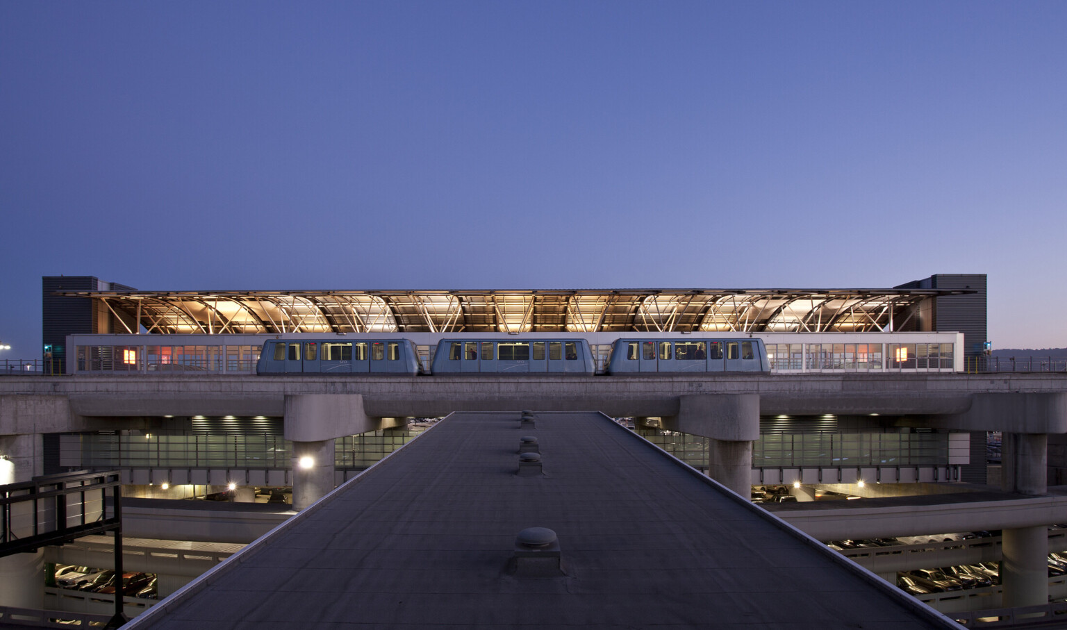 View from top of skywalk to train at station with illuminated curved half roof on opposite side with exposed beams.