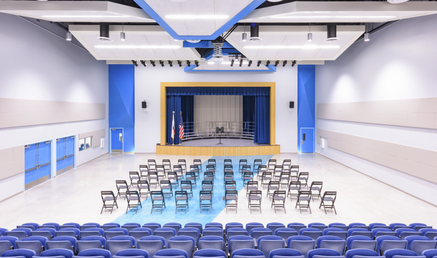 Auditorium with blue accents and doors. Folding chairs in front of blue chairs face stage with wood proscenium, blue curtains