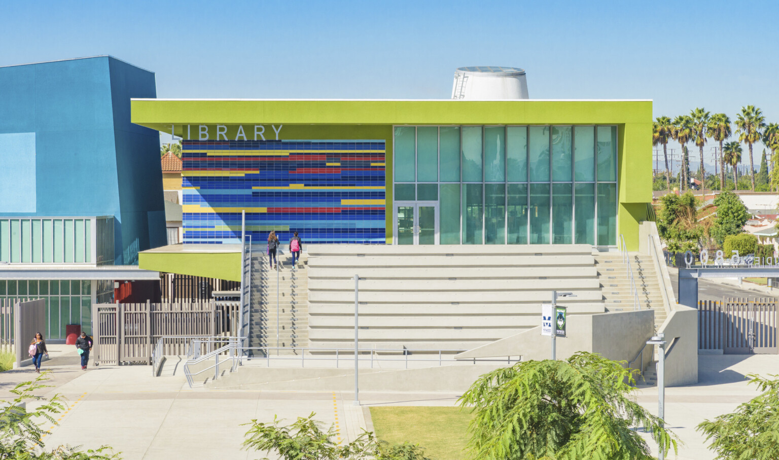 Closeup of lime green building labeled Library. Stairs on sides of step benches. Multicolor tile by recessed glass entrance