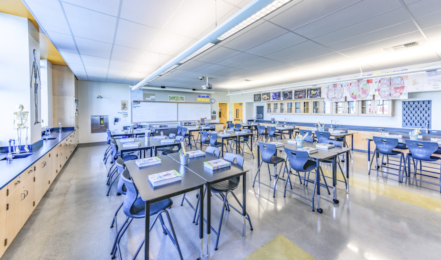 Interior classroom with flexible tables and blue chairs. Light wood cabinets with blue counters. Whiteboards on left wall
