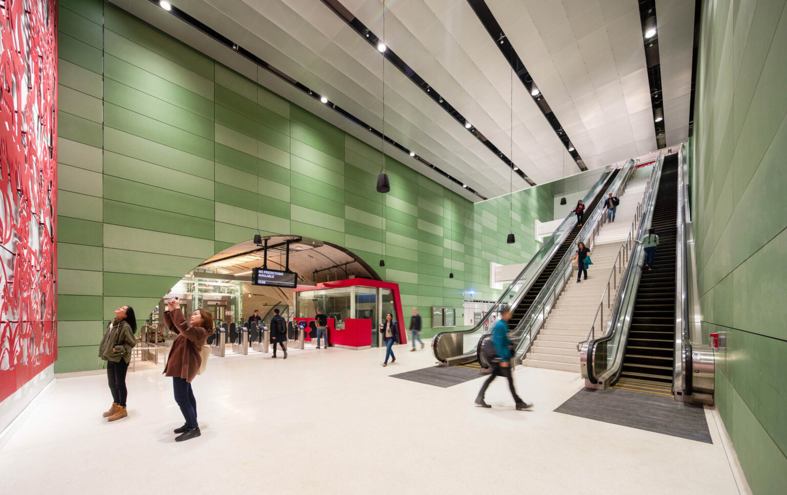 2 people looking at red art mural in a sub ground train station, escalators to back, rounded entry over turnstiles left