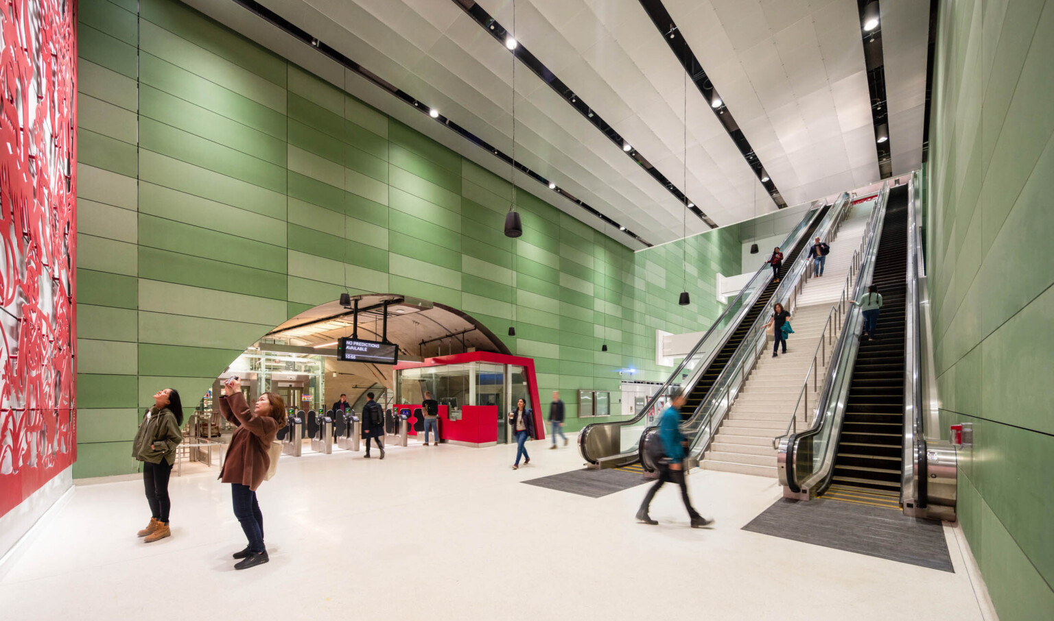 2 people looking at red art mural in a sub ground train station, escalators to back, rounded entry over turnstiles left