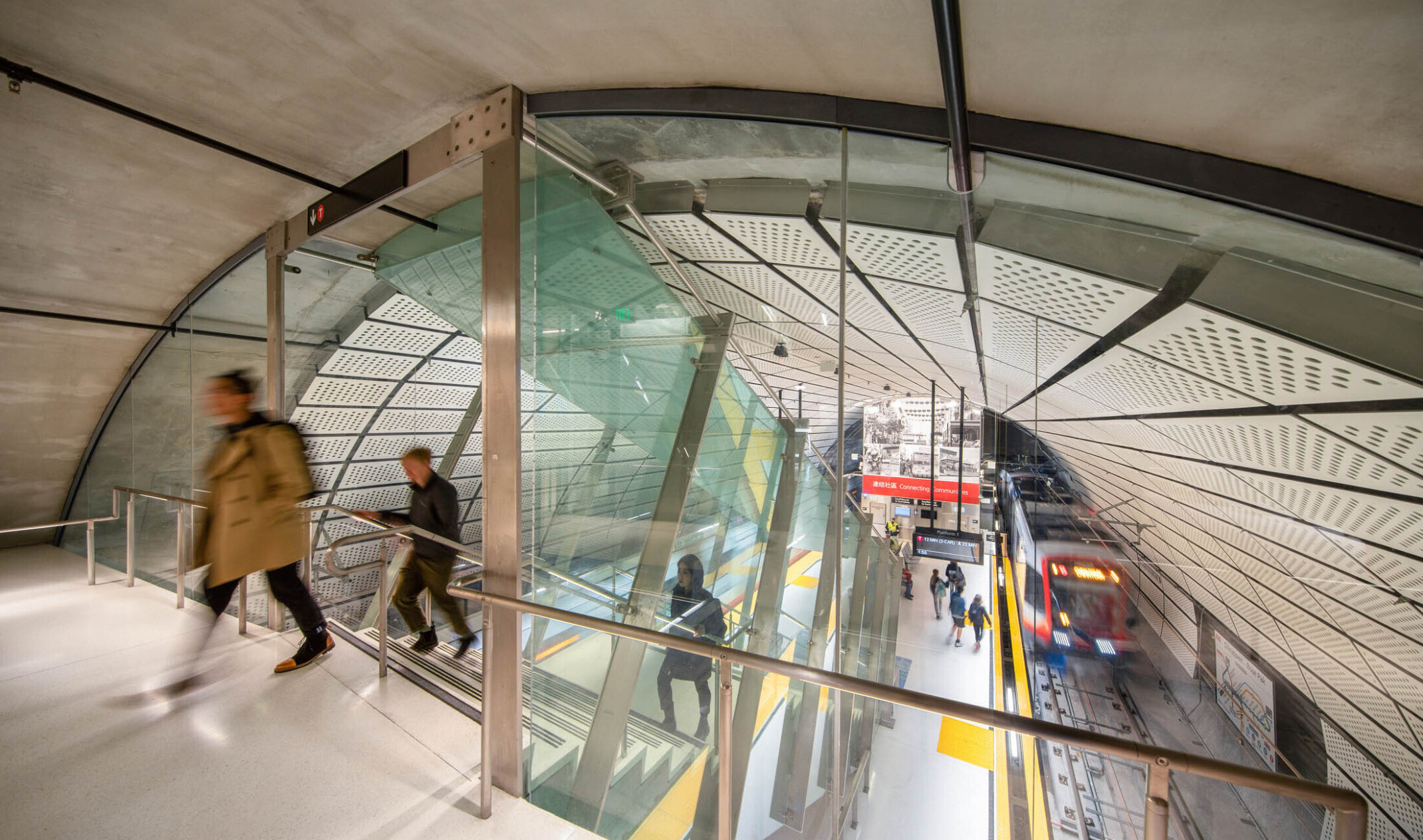 Rounded room with escalator to station below between 2 train tracks. White panel ceiling. Glass wall dividing tunnel