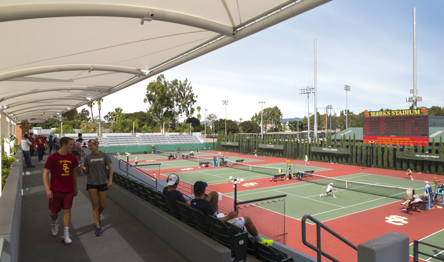 Covered walkway behind stadium seating looking to tennis courts. Tree lined far wall, scoreboard above with Marks Stadium sign
