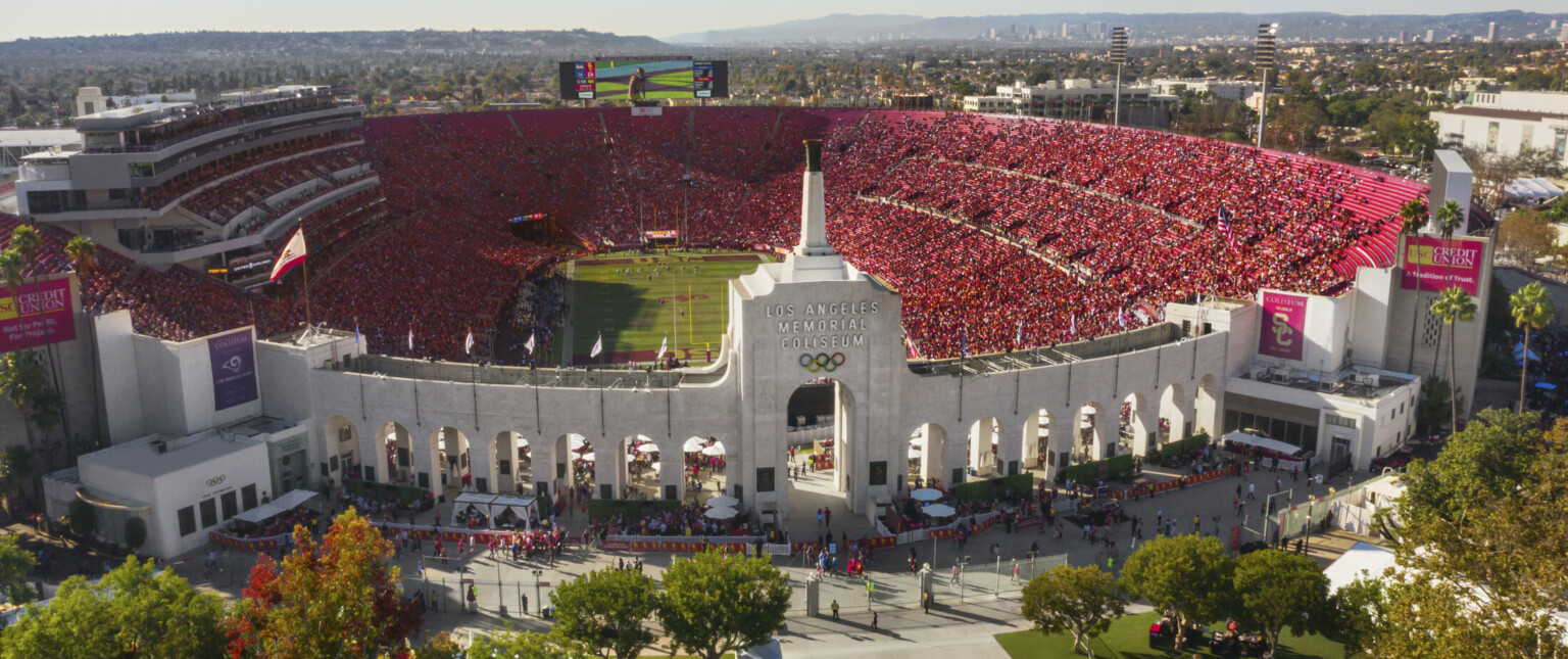 Front view of full stadium labeled Los Angeles Memorial Coliseum with Olympic Rings logo on central tower framed by archways