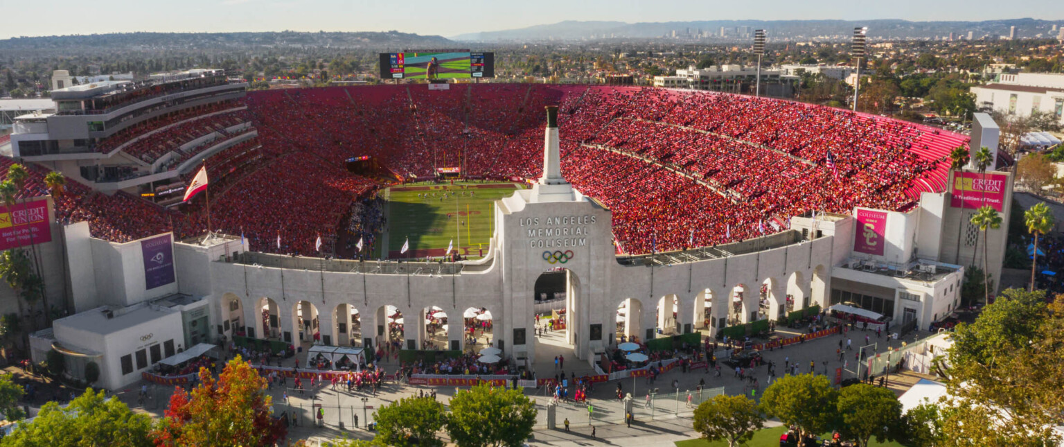 Front view of full stadium labeled Los Angeles Memorial Coliseum with Olympic Rings logo on central tower framed by archways
