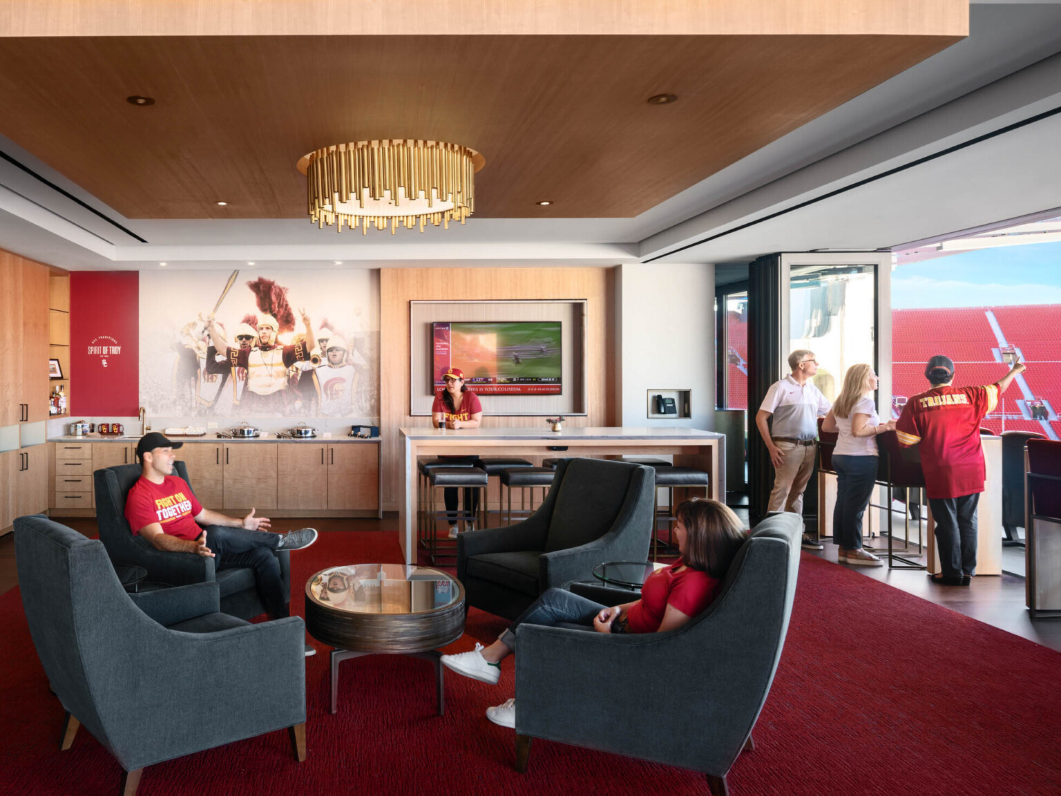 Private box in stadium with red carpet and gold chandelier. Flexible wall folds open facing field. Counter space and seating