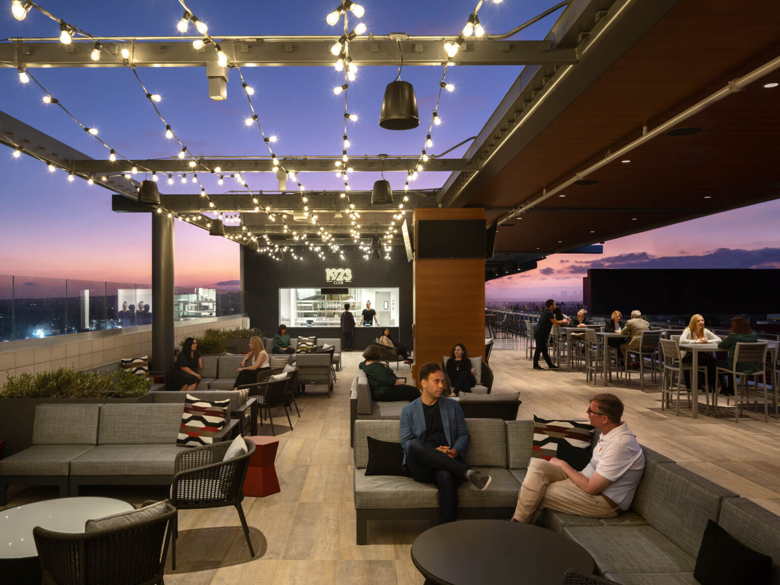 Partially covered rooftop patio with bar labeled 1923 Club. String lights hang from exposed beams at left over lounge seating