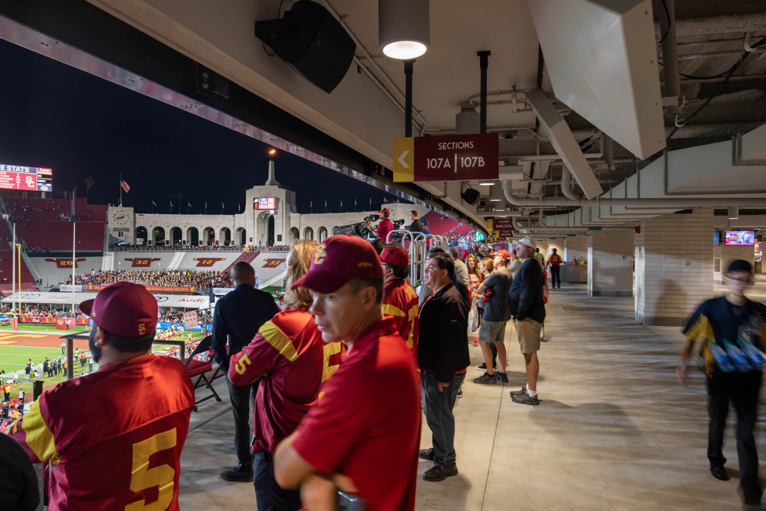 Fans stand at edge of pathway between seats and concessions area, looking at field. Exposed pipes above, brick column back