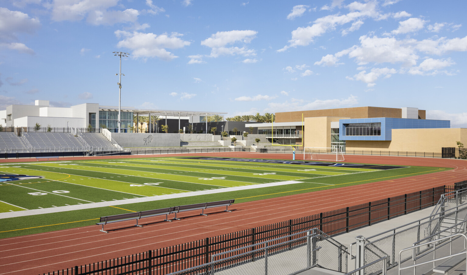 Football field surrounded by track with white bleachers, school beyond with blue accent hallway with floor to ceiling windows Football field surrounded by track with white bleachers, school beyond with blue accent hallway with floor to ceiling windows
