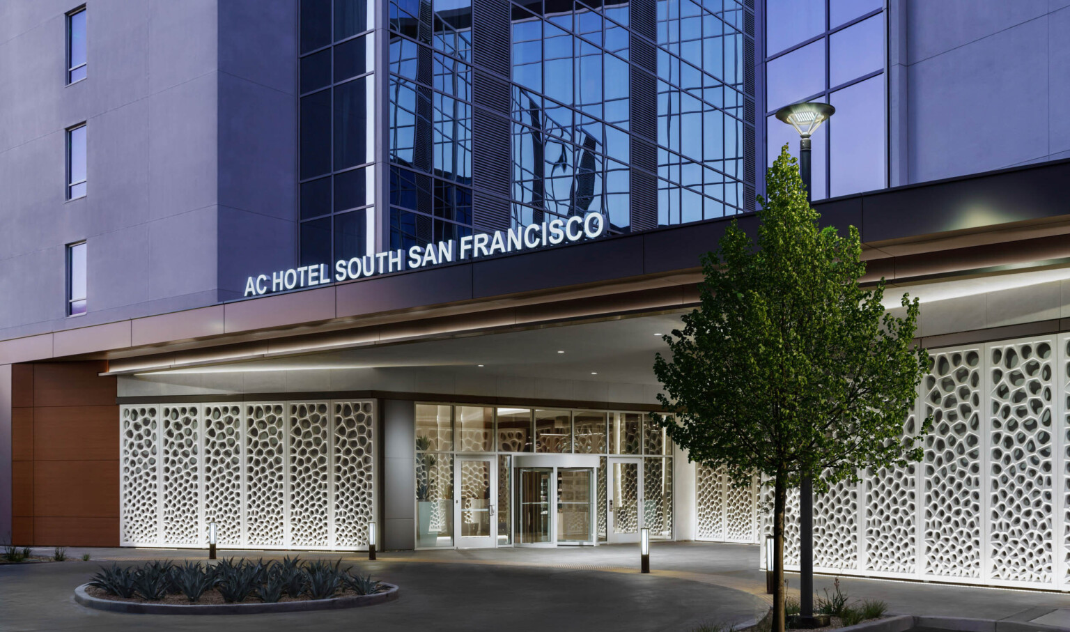 Exterior view of hotel entry with illuminated sign above. White organic sculpture lines walls under driveway shelter at entry