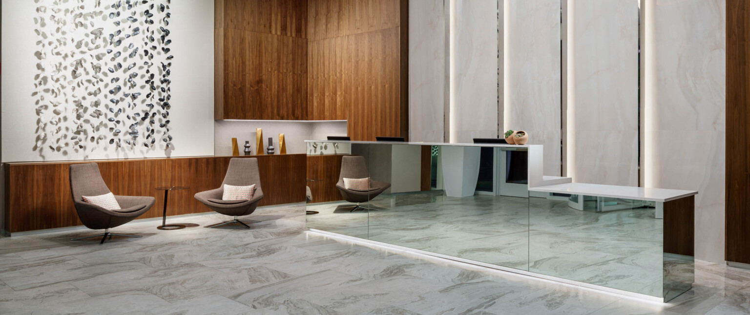 Mirrored reception desk with white counter in white lobby. Wood counter on left wraps up the wall at the corner near seating