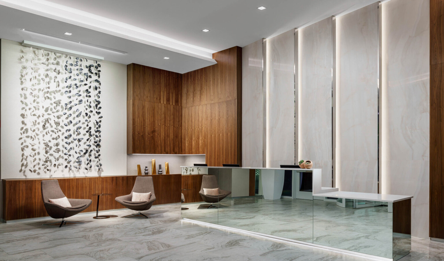Mirrored reception desk with white counter in white lobby. Wood counter on left wraps up the wall at the corner near seating