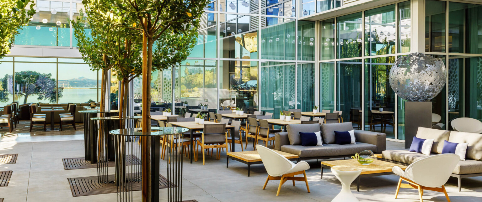 Outdoor seating with tables, chairs, couches and coffee tables lined with trees. Courtyard can be viewed from large windows following Marriott brand architecture