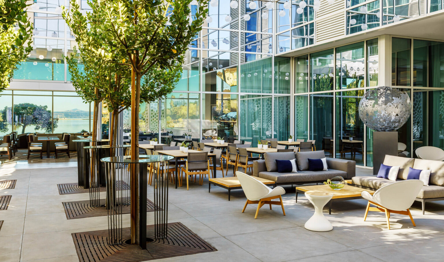 Outdoor seating with tables, chairs, couches and coffee tables lined with trees. Courtyard can be viewed from large windows