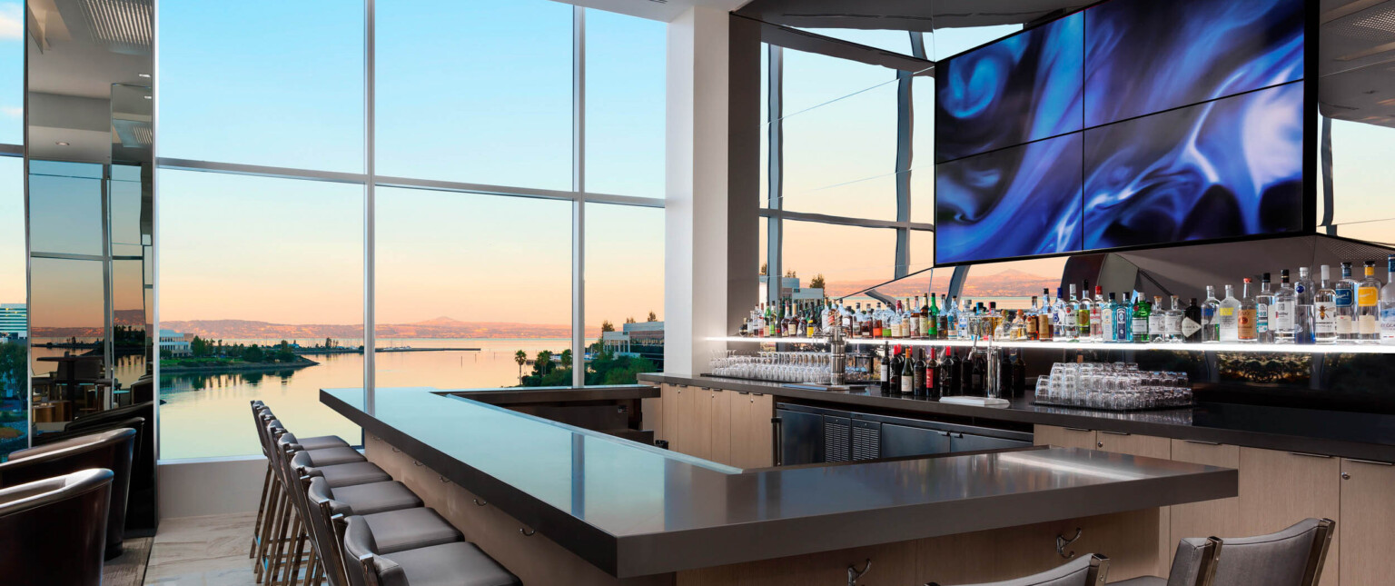 Wood bar with grey counter in room with floor to ceiling windows in front of mirror with central abstract swirling panels