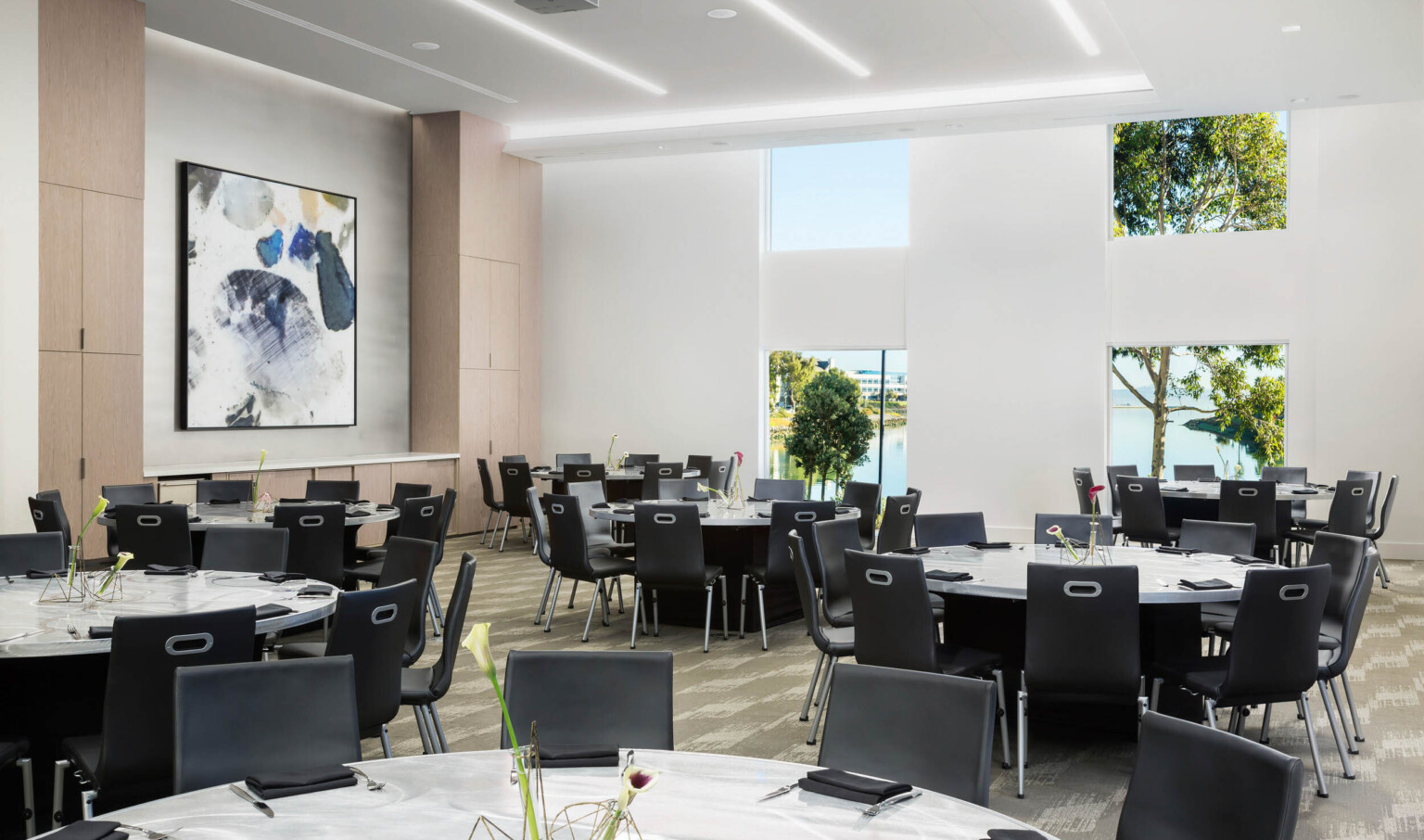 Carpeted seating area with round tables and black chairs. Wood cupboards on left wall with white counter space and framed art