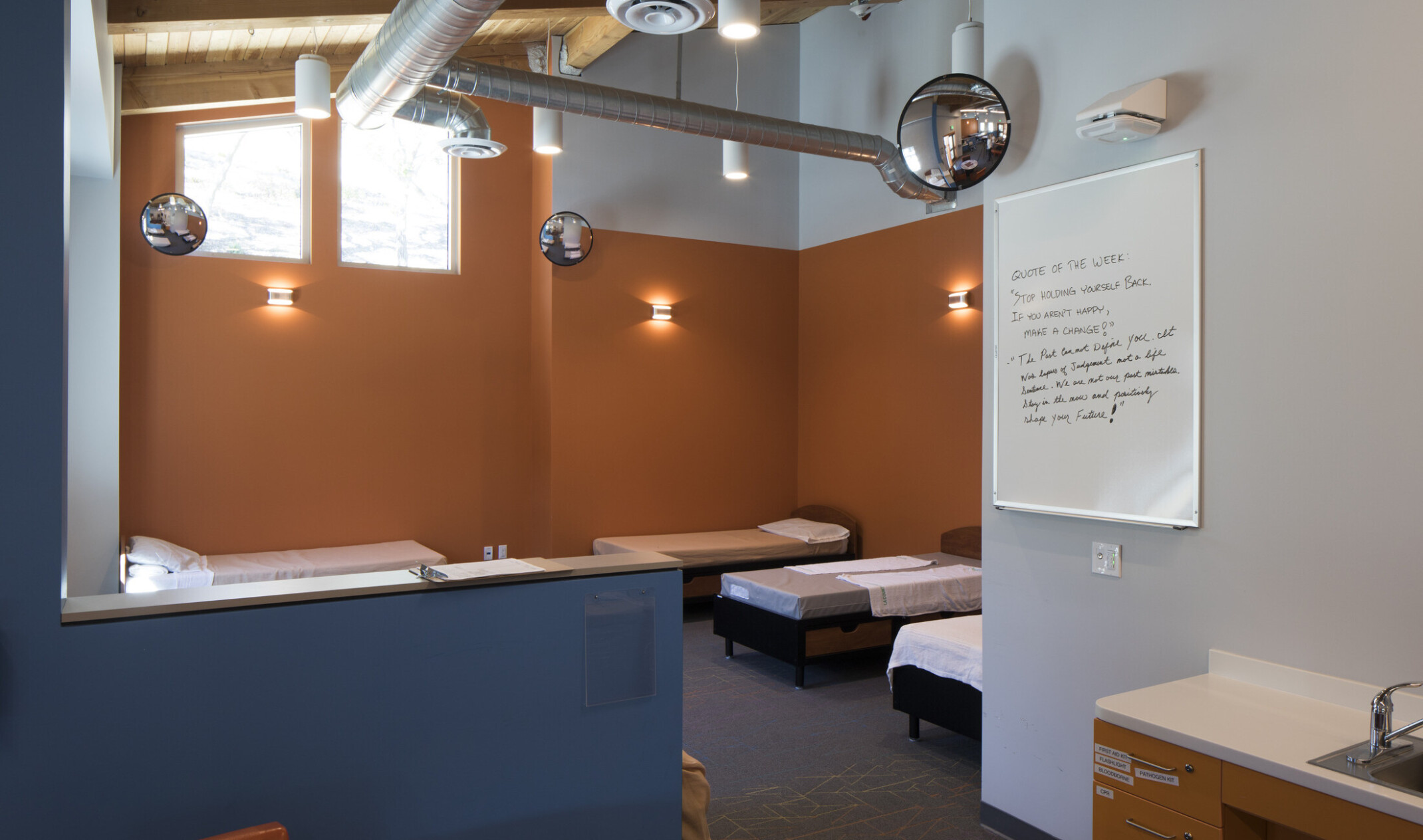 Orange bedroom with 6 beds separated from seating area by blue half wall. Exposed ductwork and round mirrors overhead