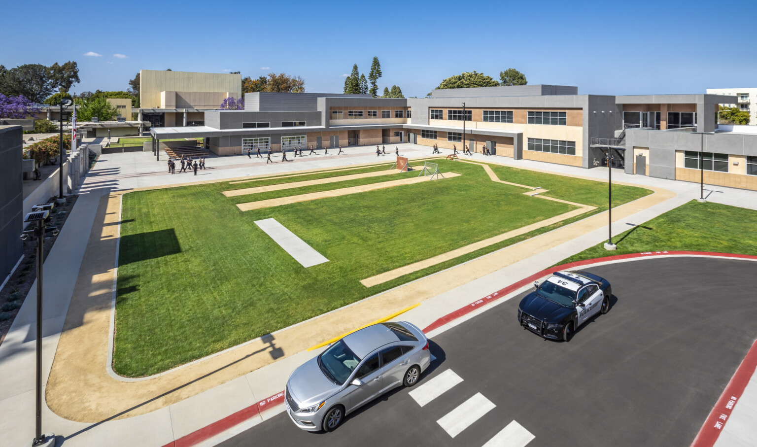 Corner aerial view of track and field with L shaped two story building in background, between which people train