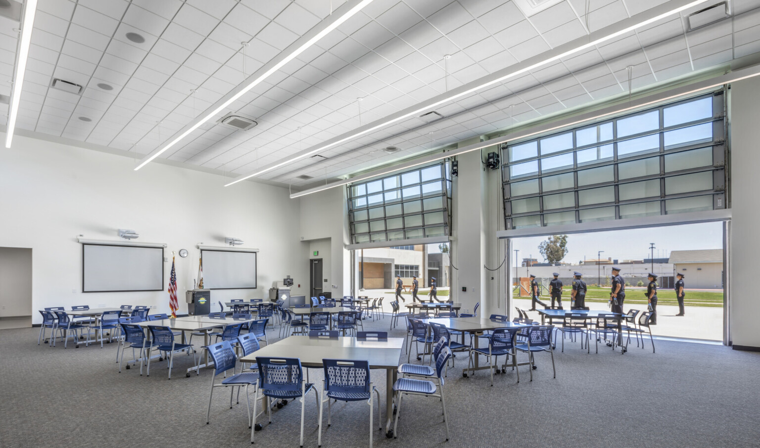 A large flexible work space with movable tables and chairs in front of a podium. Garage style doors are open to a field