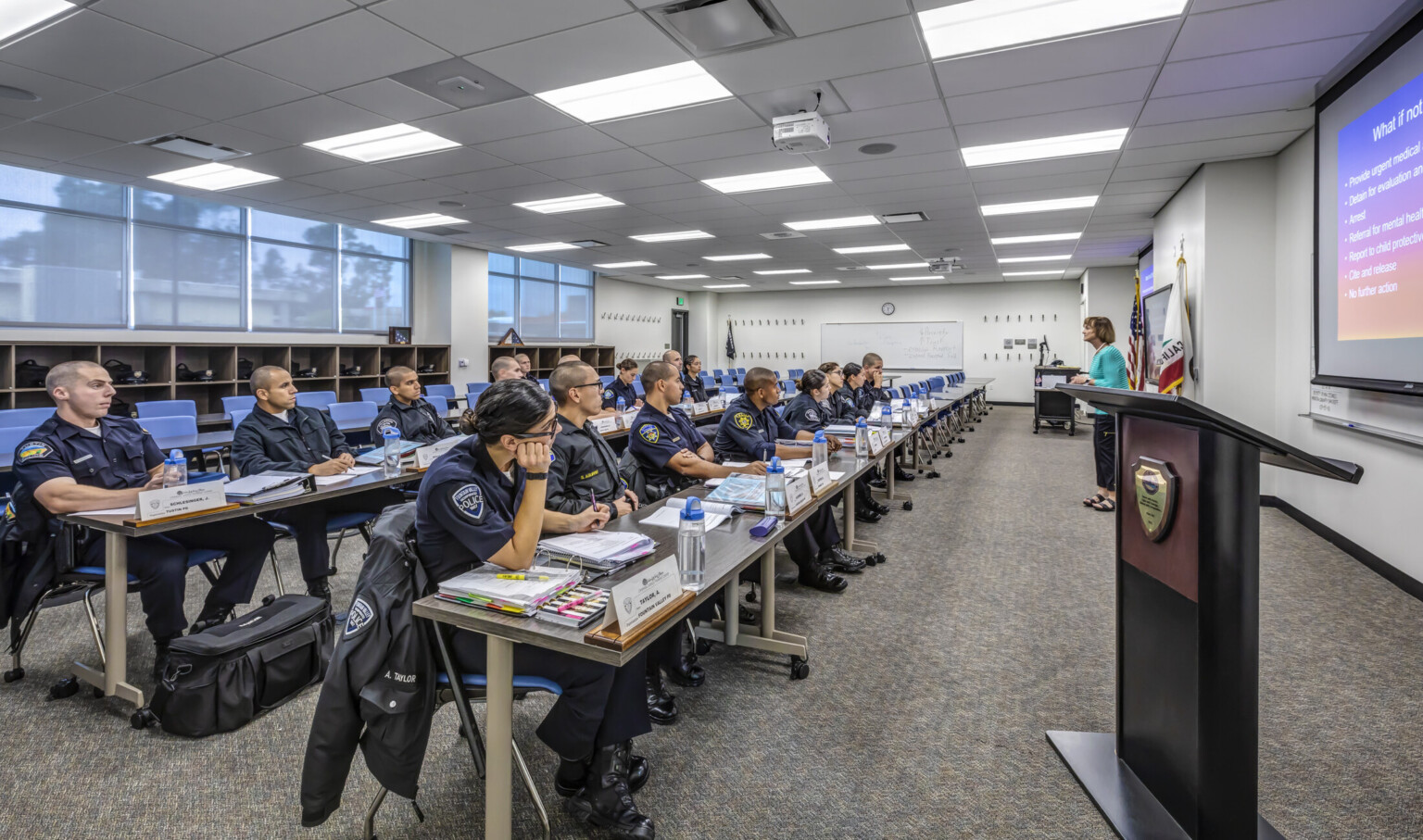 Officers sit in classroom with four long sections of rolling tables facing teacher at the front of the room
