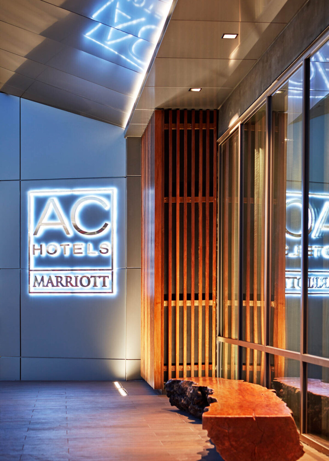 Illuminated signage on white exterior wall reads AC Hotels Marriott, beside wood panel corner detail following Marriott brand architecture