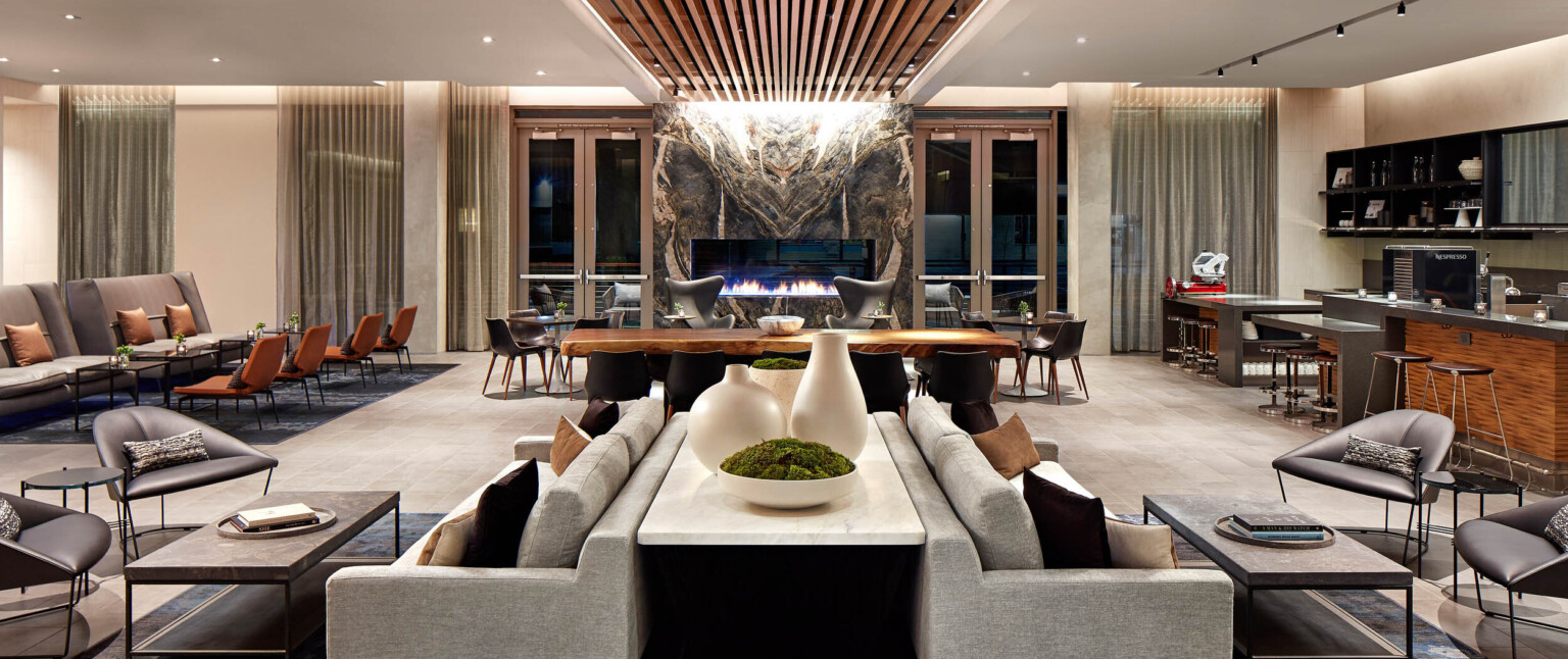 Seating area with couch and tables to the front, wooden table, and armchairs in front of organically carved stone fireplace following Marriott brand architecture