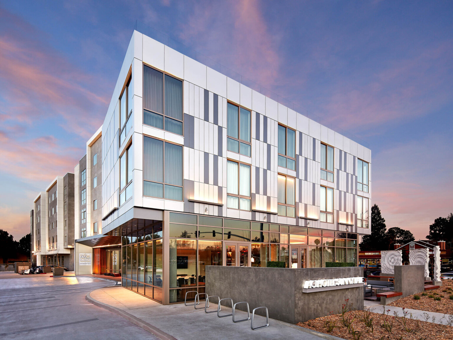 Exterior corner view of white building with recessed first story with floor to ceiling windows. Half walled entry with sign following Marriott brand architecture